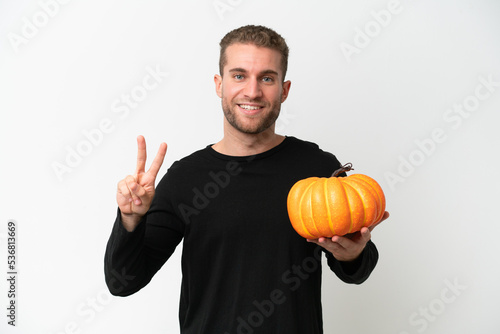 Young caucasian man holding a pumpkin isolated on white background smiling and showing victory sign © luismolinero