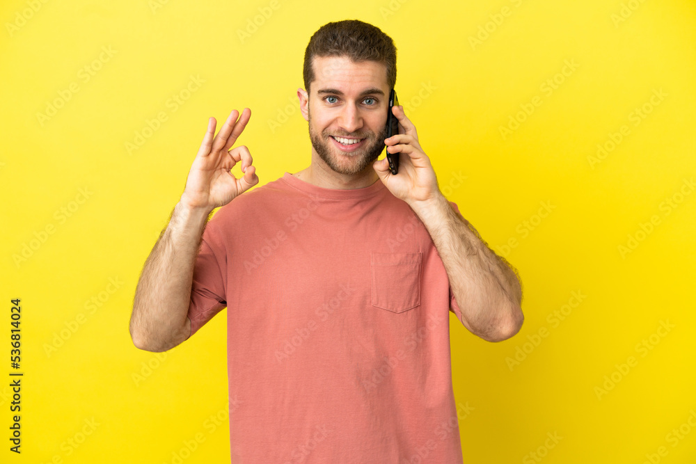 Handsome blonde man using mobile phone over isolated background showing ok sign with fingers Handsome blonde man using mobile phone over isolated background showing ok sign with fingers