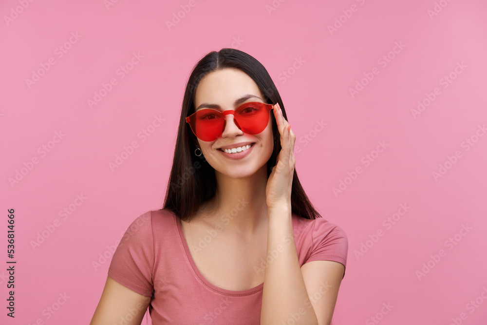 Studio close up portrait of smiling beautiful coquette young woman posing over pastel pink background in trendy red sunglasses Studio close up portrait of smiling beautiful coquette young woman posing over pastel pink background in trendy red sunglasses