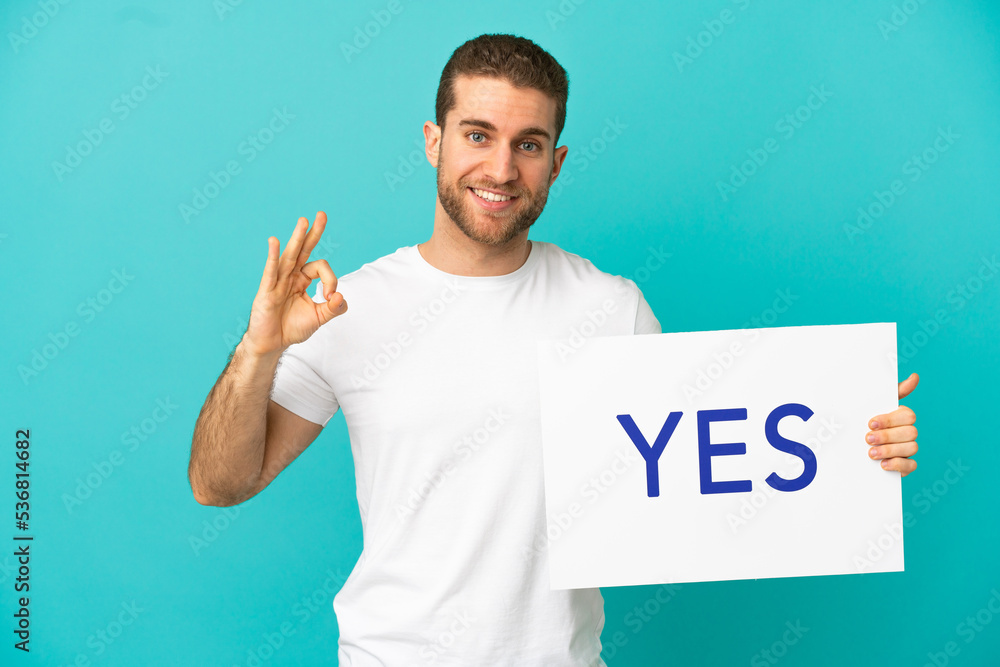 Handsome blonde man over isolated blue background holding a placard with text YES Handsome blonde man over isolated blue background holding a placard with text YES
