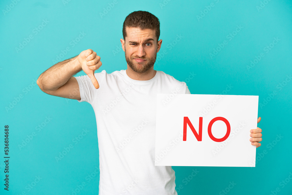 Handsome blonde man over isolated blue background holding a placard with text NO and doing bad signal Handsome blonde man over isolated blue background holding a placard with text NO and doing bad signal