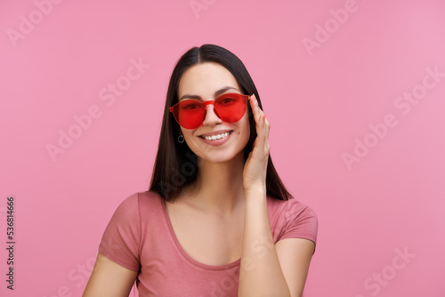 Studio close up portrait of smiling beautiful coquette young woman posing over pastel pink background in trendy red sunglasses