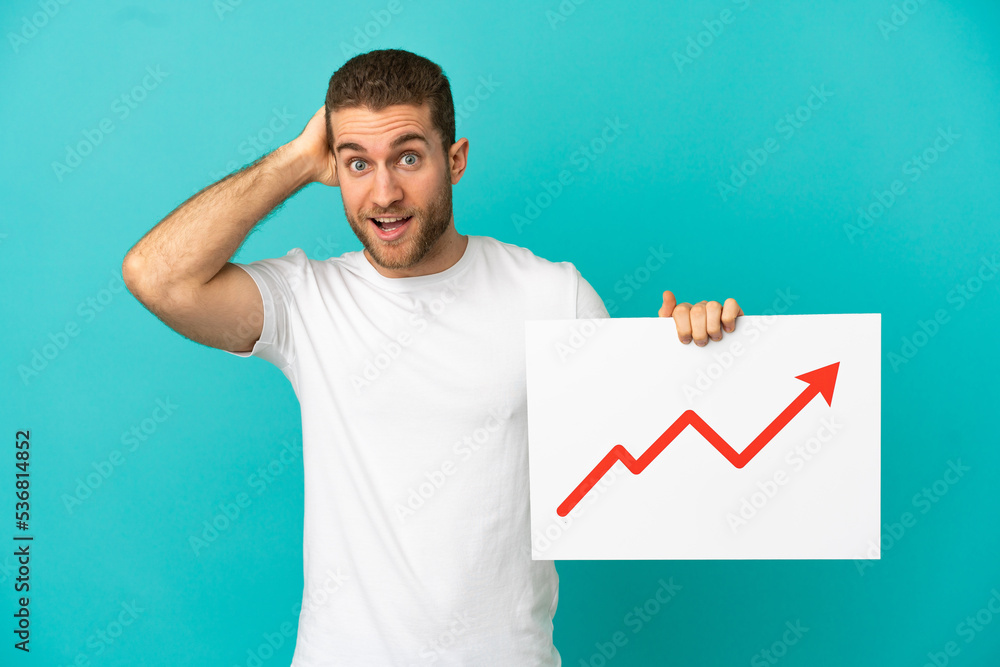 Handsome blonde man over isolated blue background holding a sign with a growing statistics arrow symbol with surprised expression Handsome blonde man over isolated blue background holding a sign with a growing statistics arrow symbol with surprised expression