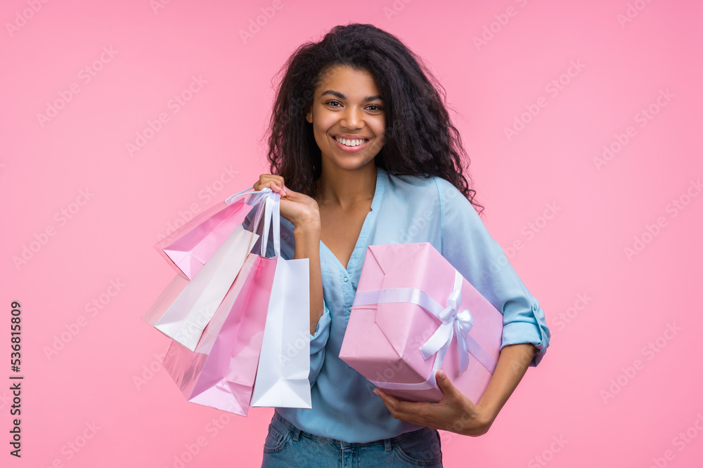 Studio portrait of beautiful happy smiling birthday girl holding decorated gift box and a bunch of shopping bags in hands, isolated over pastel pink background Studio portrait of beautiful happy smiling birthday girl holding decorated gift box and a bunch of shopping bags in hands, isolated over pastel pink background