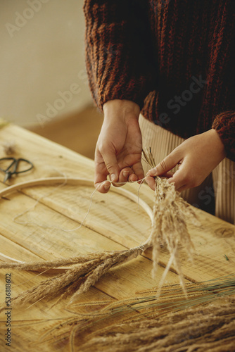 Making stylish autumn wreath on rustic table. Hands arranging dried grass on hoop on wooden table with scissors, thread. Fall decor and arrangement in farmhouse.