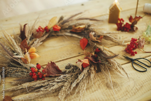 Stylish autumn wreath on rustic table. Rustic autumn wreath with dried grass, berries, herbs on wooden table with candle, scissors. Fall decor and arrangement in farmhouse. Thanksgiving