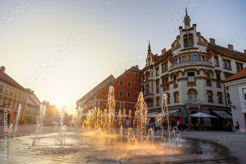 Splashing fountain on square in Maribor with backlit. Historical building in center of the city in Slovenia with streaming water in golden hour. Old street with flowing waterwork in sunset.
