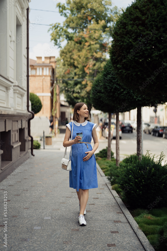 Beautiful young pregnant woman in a blue dress walking through the city streets having a take away drink.