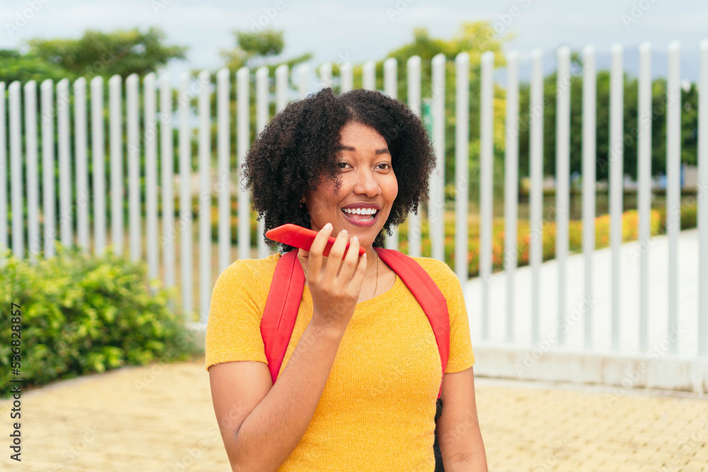 Fototapeta premium Smiling woman sending a voice message via cell phone in the park.