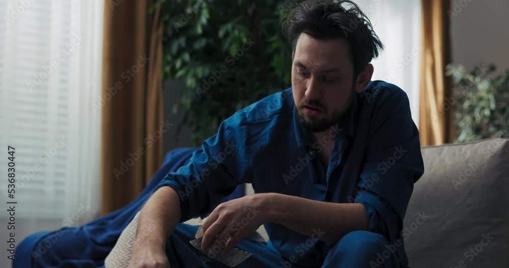 Close-up of man in blue pajamas sitting on bed and taking pills. He has fever. He holds the pills in hands and chooses the right one. The man press the pill on hand and drinks it with tea. Close-up of man in blue pajamas sitting on bed and taking pills. He has fever. He holds the pills in hands and chooses the right one. The man press the pill on hand and drinks it with tea.