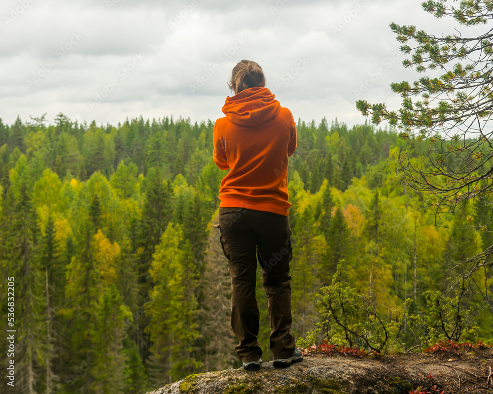 Naklejka premium a woman in a hoodie and trousers stands on the edge of a cliff against the background of a forest. the concept of travel and tourism