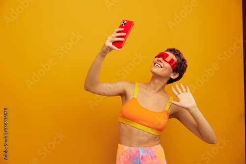 Woman blogger smiling in unusual millennial glasses taking selfies on her phone in sportswear against an orange studio backdrop  free space