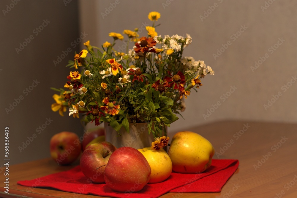 Autumn still life, bouquet of flowers on the table, in the foreground ripe apples on a red cloth