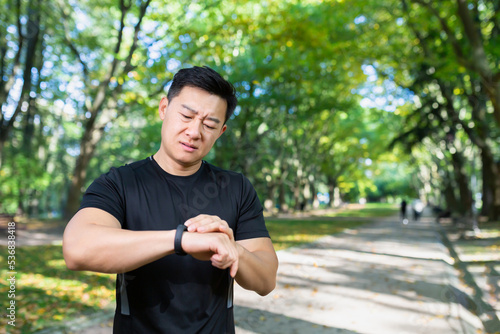 unhappy and upset male athlete looking at fitness with smartwatch, bad workout result, asian man exercising in autumn park on day.