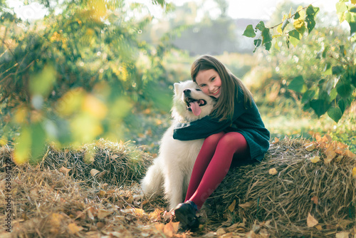 A little brunette girl with her white fluffy dog lies on autumn leaves in the park. Friendship and harmony in the family. Happy childhood.
