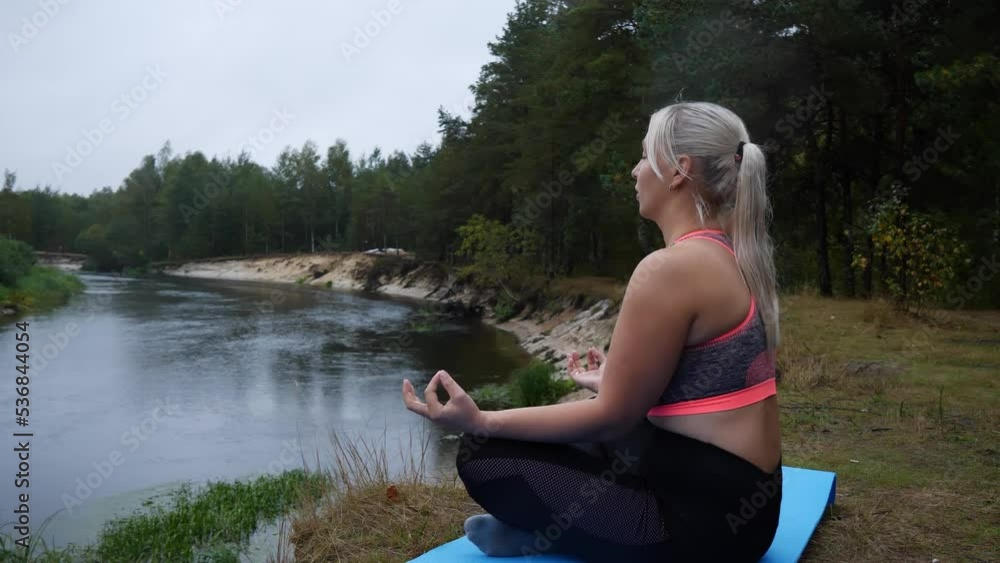 In the lotus position on the edge of a steep river bank, a woman breathes slowly with her eyes closed. A young woman practices yoga and meditation on a beautiful river bank. Breath concept. In the lotus position on the edge of a steep river bank, a woman breathes slowly with her eyes closed. A young woman practices yoga and meditation on a beautiful river bank. Breath concept.