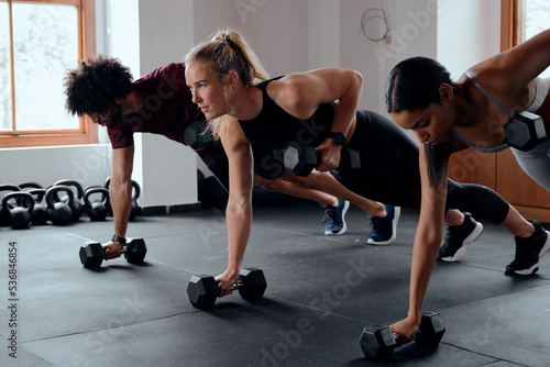 Three focused young adults in a row doing plank exercises with weights at the gym