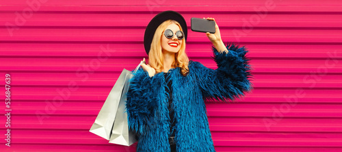 Portrait of happy smiling blonde woman taking selfie with smartphone with shopping bags wearing blue fur coat, black round hat on pink background © rohappy