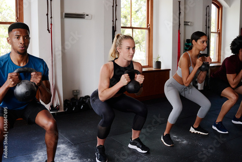 Group of multiracial young adults in a row doing kettlebell squats at the gym