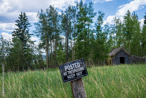 field with tall grass and old wooden building in the background with focus on sign that says Posted No Trespassing Keep Out