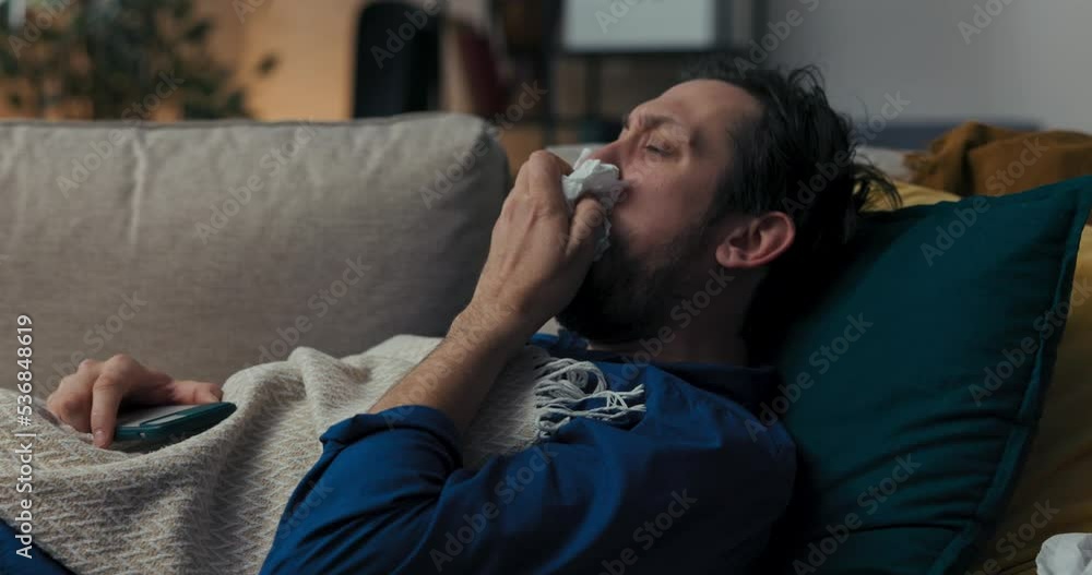 Sick man with beard is lying on sofa on blue pillow and covered with blanket. He does not feel well, picks up napkin and coughs loudly. Brunette is holding the phone and scrolling through the news. Sick man with beard is lying on sofa on blue pillow and covered with blanket. He does not feel well, picks up napkin and coughs loudly. Brunette is holding the phone and scrolling through the news.