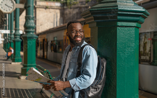 Happy young african american man waiting with map at european terminal train station. Tourist travel by train on vacation time holiday weekend trip.
