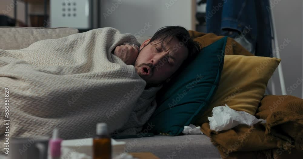 Sick man is lying on sofa, covered with blanket. He is not feeling well and is sleeping. The man coughed hard, woke up and looked at the time on phone. There are medicines and napkins on the table. Sick man is lying on sofa, covered with blanket. He is not feeling well and is sleeping. The man coughed hard, woke up and looked at the time on phone. There are medicines and napkins on the table.