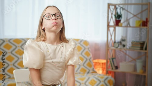 Stylish girl making funny face. Girl in stylish blouse and glasses with long hair puffing and touching cheeks while sitting on chair near couch in living room at home.
