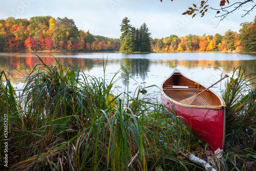 Fototapeta Naklejka Na Ścianę i Meble -  Red wooden canoe on shore of lake with trees in autumn color and a small island