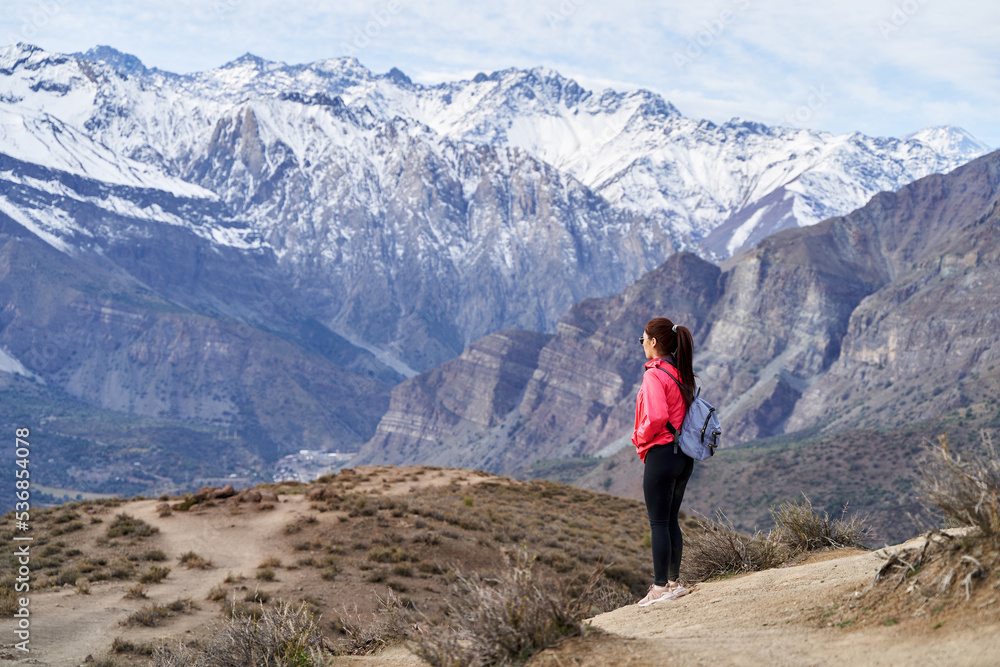 young red haired woman with red jacket and backpack, looking at the snow, in the middle of the Andes mountain range in chile