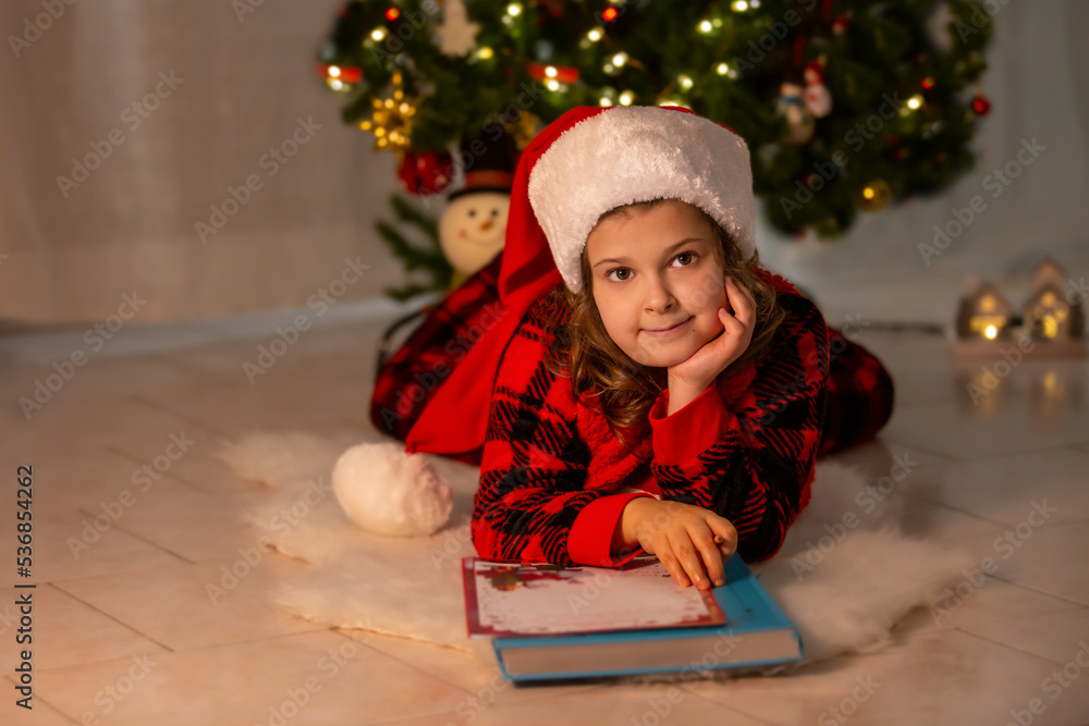 A little girl in a Santa hat writes a letter to Santa at home lying on the bokeh floor in the background. Winter clothes for Christmas. Merry Christmas and Happy Holidays A little girl in a Santa hat writes a letter to Santa at home lying on the bokeh floor in the background. Winter clothes for Christmas. Merry Christmas and Happy Holidays