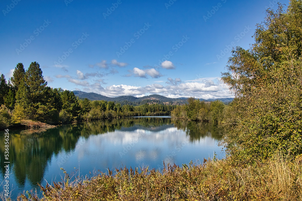 On a sunny Autumn day in Idaho, a peaceful river with a glassy surface reflects the sky and surrounding mountainous landscape.