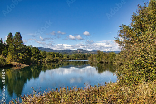 On a sunny Autumn day in Northwestern Idaho, a peaceful, reflecting mountain river is viewed from the Trail of the Coeur d'Alenes, with clouds and mountains in the background.