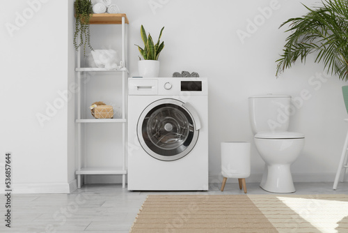 Modern washing machine, shelf unit and toilet bowl near white wall in bathroom interior © Pixel-Shot