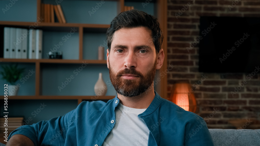 Head shot handsome bearded caucasian guy confident serious 30s male portrait wears casual blue shirt attractive caucasian man homeowner client sit on sofa at home looking at camera with serene emotion Head shot handsome bearded caucasian guy confident serious 30s male portrait wears casual blue shirt attractive caucasian man homeowner client sit on sofa at home looking at camera with serene emotion
