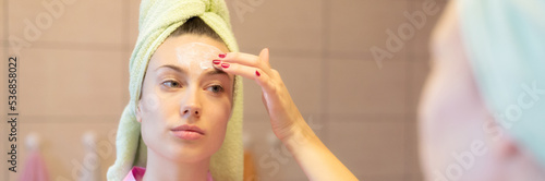 Young woman putting facial mask on her face in front of the bathroom mirror