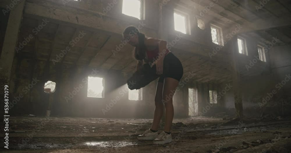 A young woman in fitness clothes lifts a heavy railway sleeper as part of physical training in an old industrial hall. Low angle wide shot. A young woman in fitness clothes lifts a heavy railway sleeper as part of physical training in an old industrial hall. Low angle wide shot.