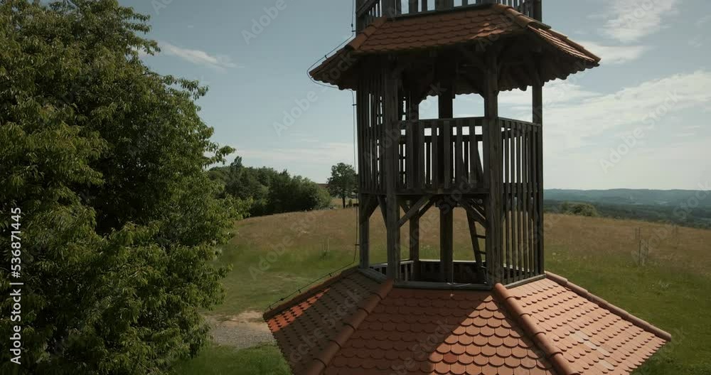 Aerial panoramic drone shot of a hiker man with backpack who rings a bell on a top of a small wooden tower in nature with green forest and grass agricultural fields around the hill in Slovenia Aerial panoramic drone shot of a hiker man with backpack who rings a bell on a top of a small wooden tower in nature with green forest and grass agricultural fields around the hill in Slovenia