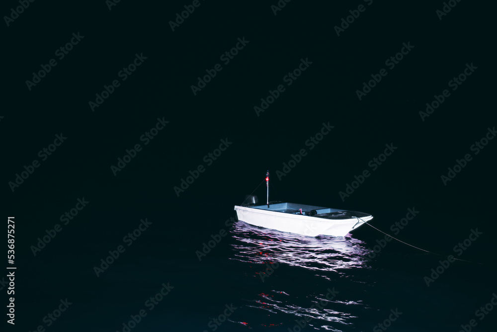 Naklejka premium A low-key shot of a small lonely moored boat in the darkness of the night illuminated by a spotlight, with a red beacon light and purple reflections in the water; a copy space area on the left