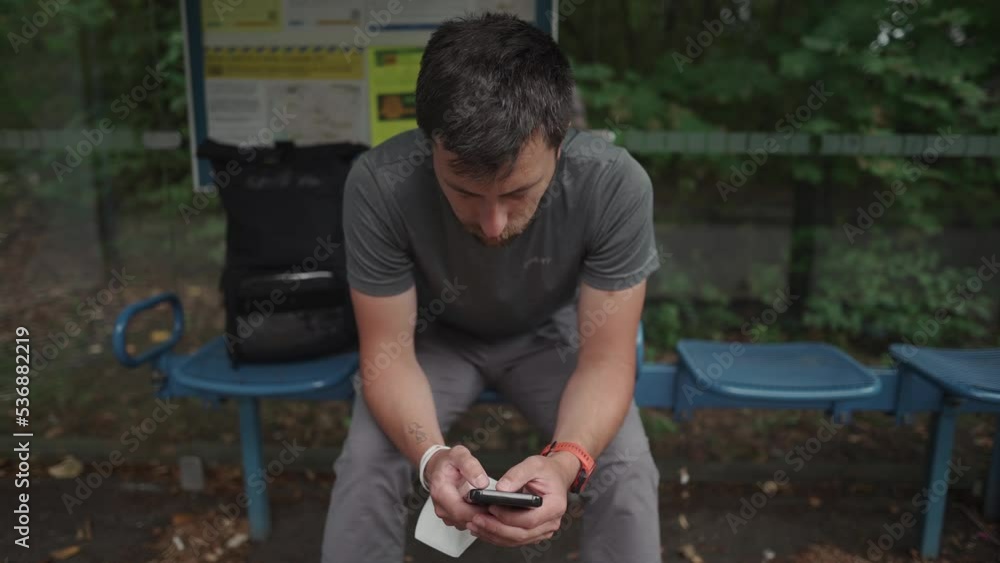 Man sits at bus stop, waiting for transport and surfing Internet on smartphone in Munich, Germany. Passenger at station texting, chatting, browsing Internet, checking email. Social media, networking. 