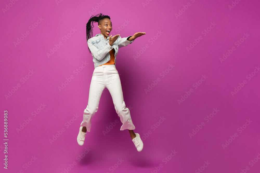 Energetic trendy young African American woman jumping and pointing sideways at copy space in studio pink color isolated background