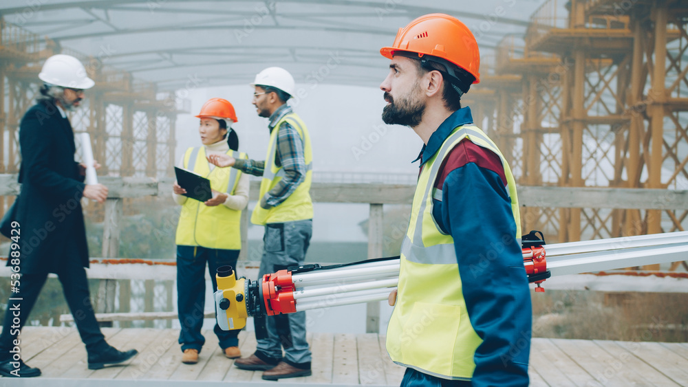 serious male surveyor in uniform walking in construction site holding theodolite concentrated on work. People and profession concept. serious male surveyor in uniform walking in construction site holding theodolite concentrated on work. People and profession concept.