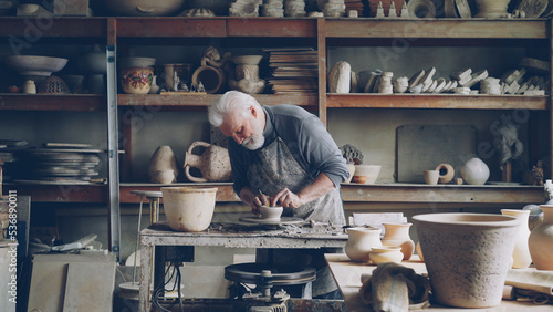 Fotografie Experienced male ceramist is creating utensils from clay on potter's wheel in workshop