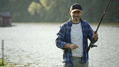 Fisherman stands on bank and unhooks bait folding rod. Fisherman stands on river bank and holds fishing rod in hands on sunny summer day. Bearded man unhooks bait and looks smiling and begins to fold photo