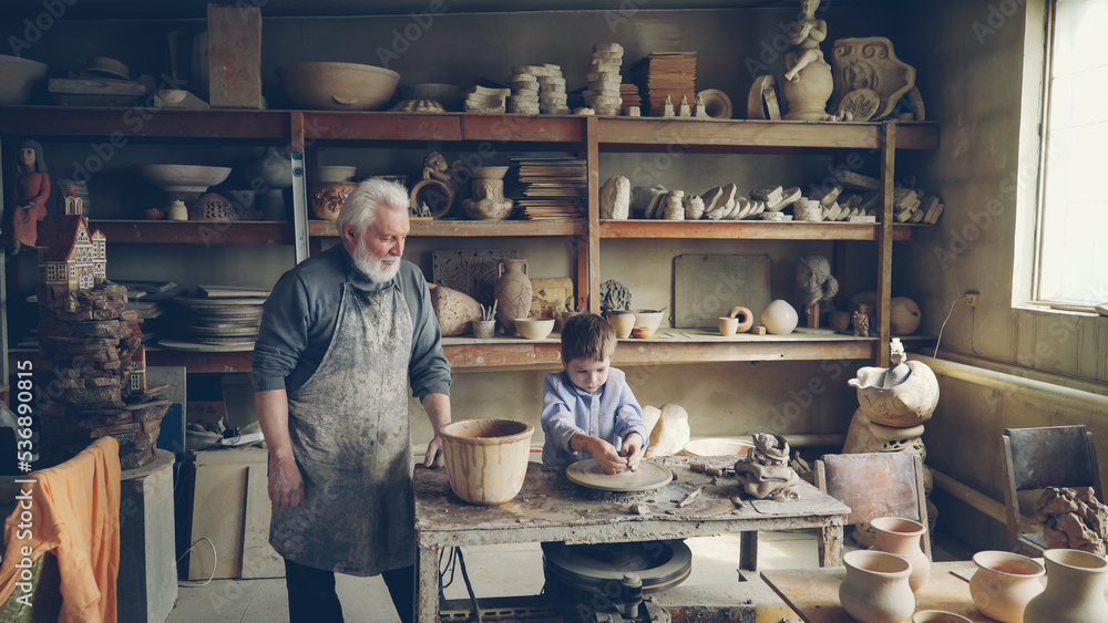 Young grandson is molding clayware in his grandfathers's home studio while his loving senior grandpa is standing near him and watching him with love. Family traditions concept. Young grandson is molding clayware in his grandfathers's home studio while his loving senior grandpa is standing near him and watching him with love. Family traditions concept.