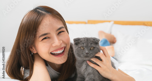 Young Asian woman lying on bed and playing small Scottish kitten on the room.