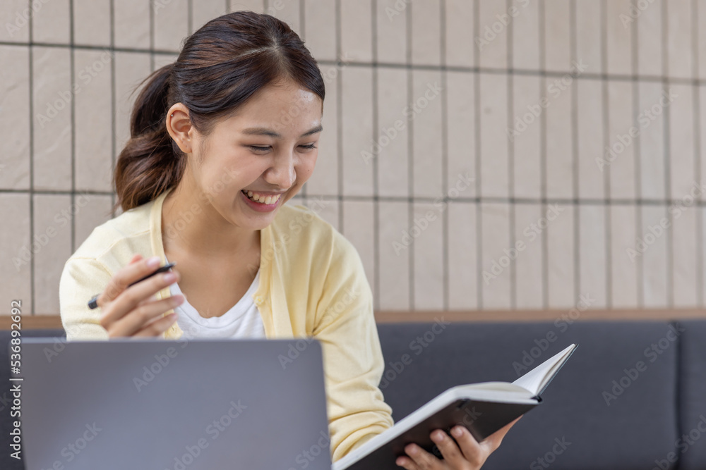 Young Asian lady studying online from home, reading a book and using laptop pc, sitting on the coffee shop, free space. E-learning, web-based education concept Young Asian lady studying online from home, reading a book and using laptop pc, sitting on the coffee shop, free space. E-learning, web-based education concept