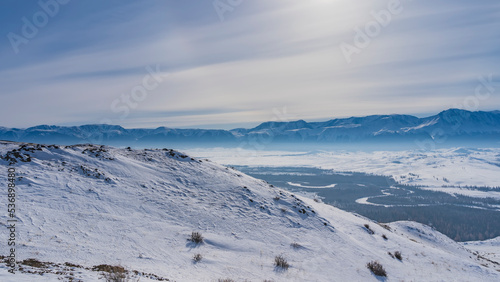 The vast expanses are visible from the top of the snow-covered plateau. Down in the valley  the river winds through the forest. A mountain range against the sky. Altai