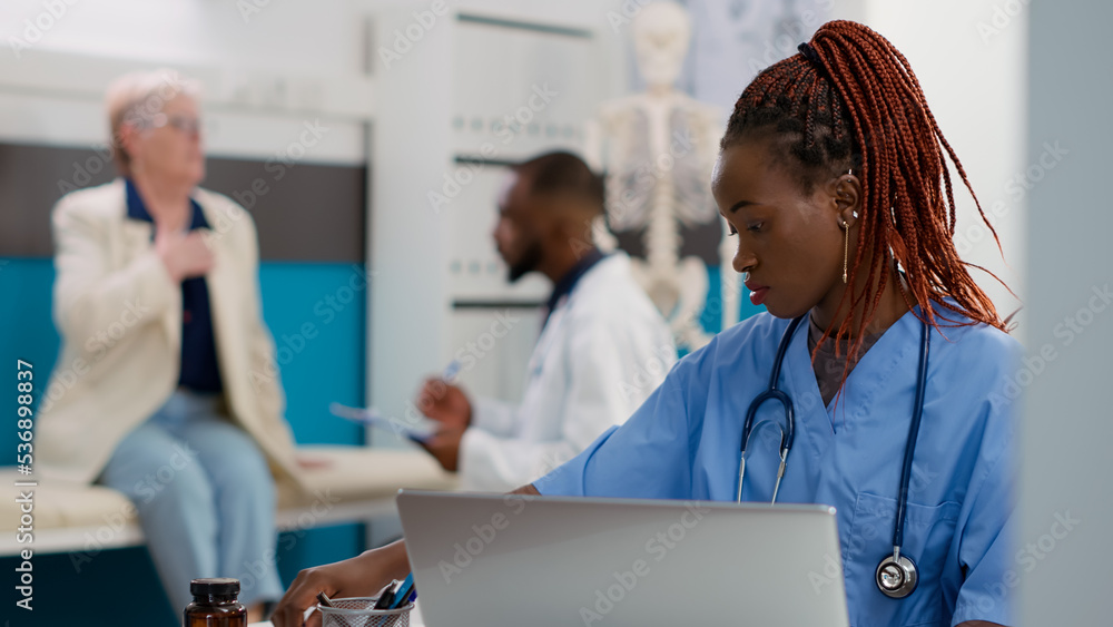 Fototapeta premium African american nurse working on appointments in cabinet, using laptop to plan checkup visits to help patients at healthcare facility. Medical assistant with stethoscope planning consultations.