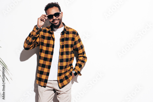 Handsome confident hipster model. Sexy unshaven man dressed in yellow summer shirt and jeans clothes. Fashion male posing near white wall in studio at sunny day. Isolated. In sunglasses